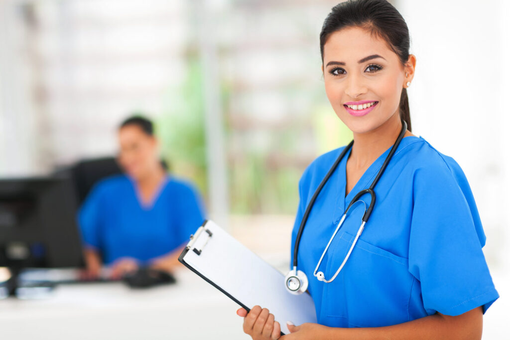 A nursing student wearing scrubs and a stethoscope, holding a clipboard, smiling at the camera.