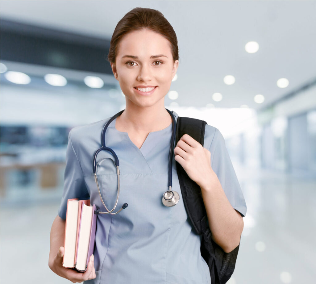 A young nursing student wearing scrubs and a stethoscope, holding school books, smiling at the camera.